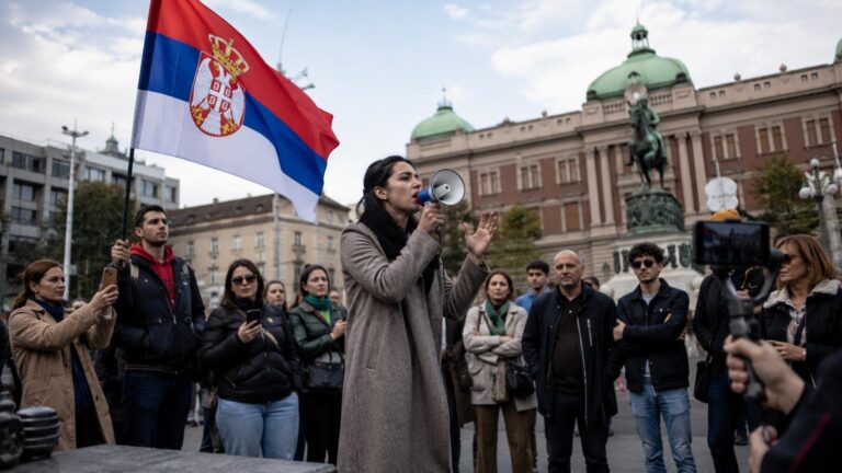 Žena govori preko megafona pred okupljenim građanima, sa zastavom Srbije i zgradom institucije u pozadini